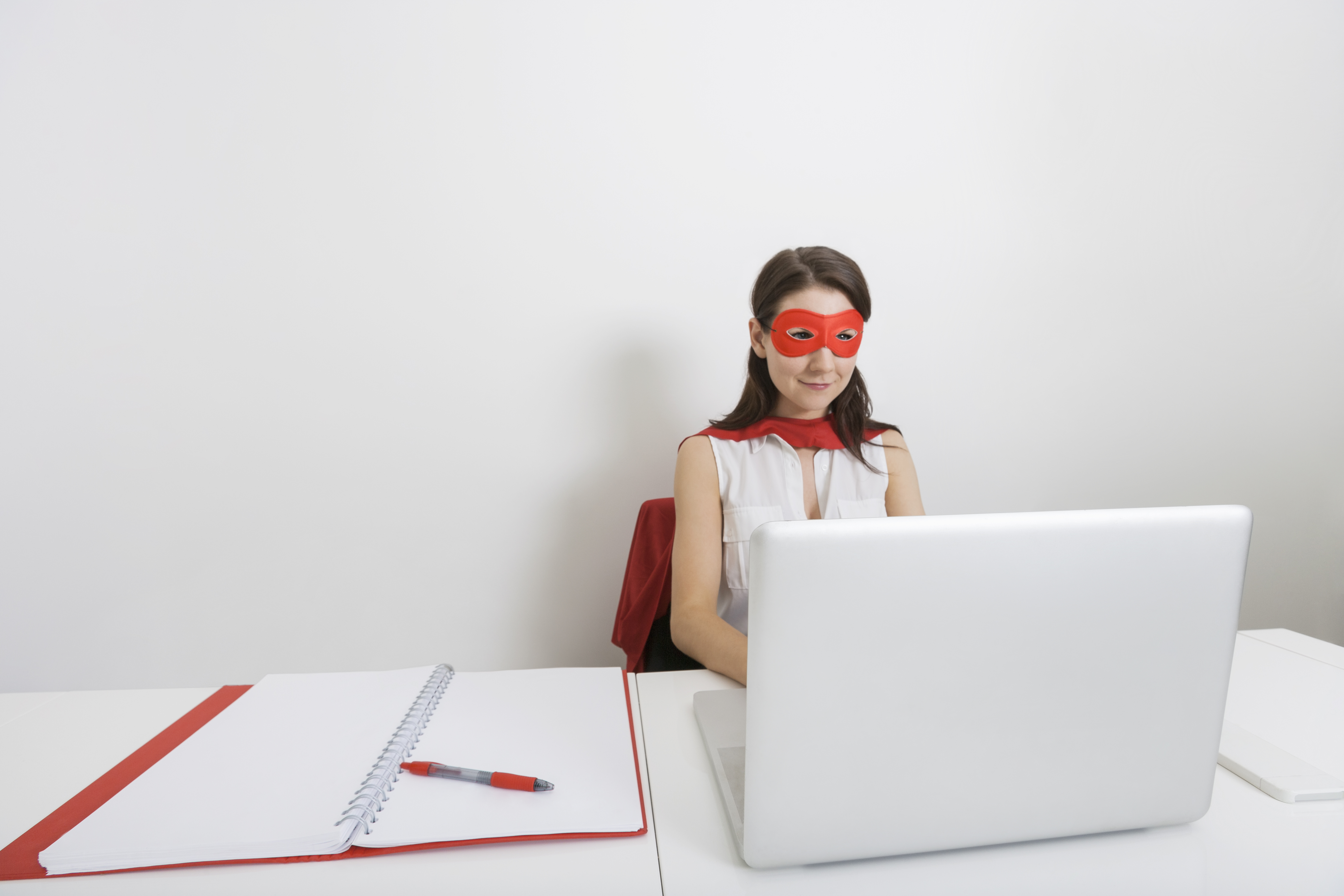 Young woman wearing red eye mask and focusing on white computer with notebook and pen beside her