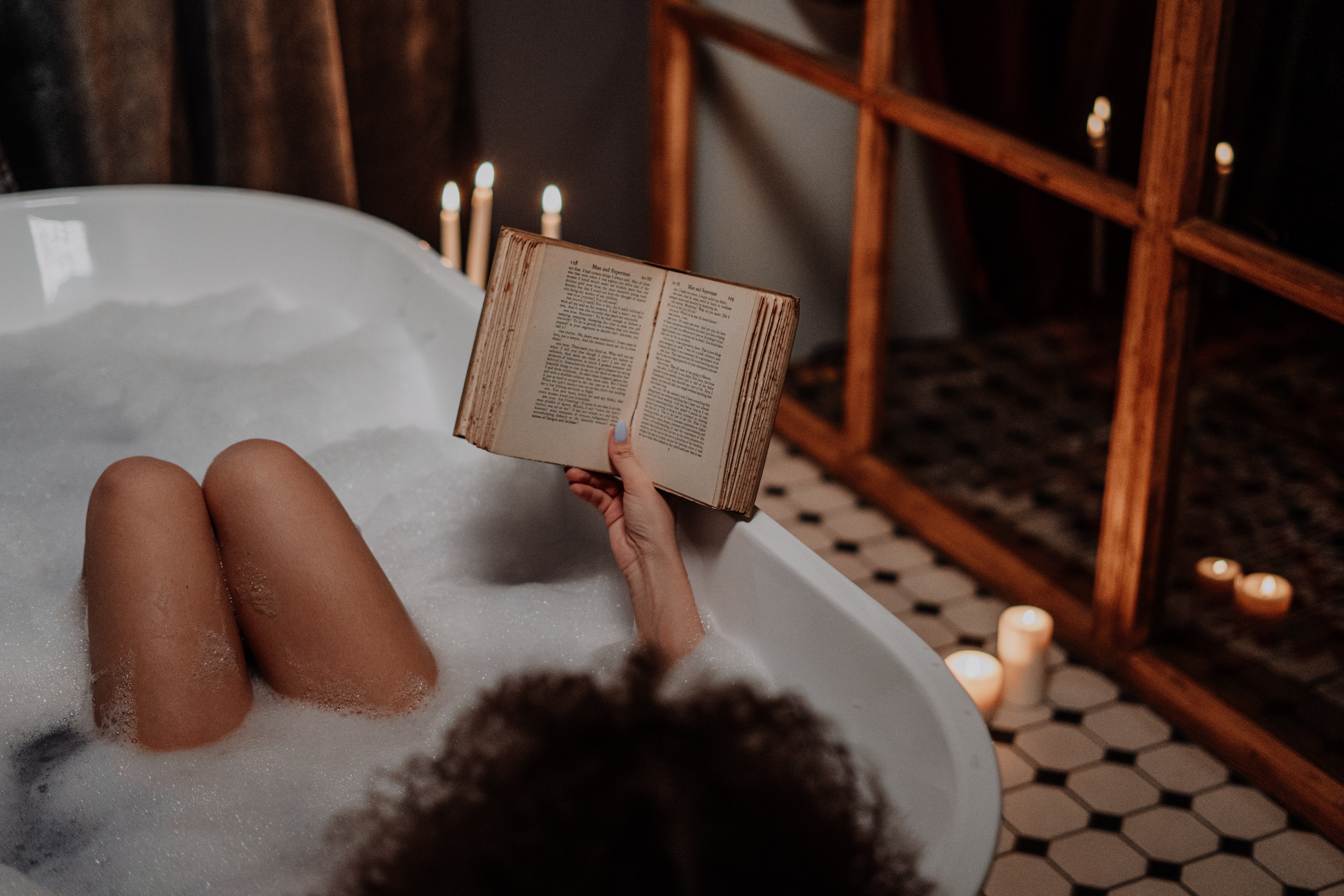 Woman having a bath in a white bathtub reading a book surrounded by candles