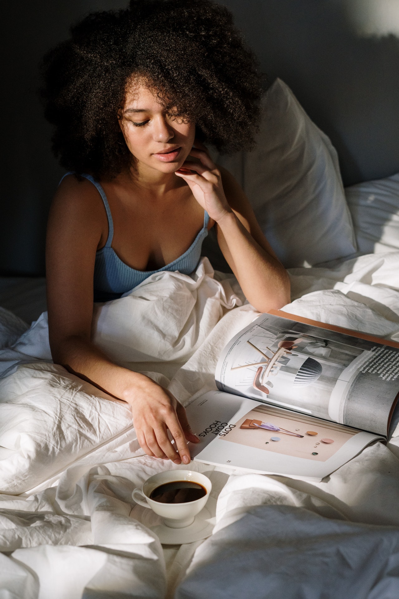 Girl with curly, black hair reading magazine with a cup of coffee in a bed in relaxing mood 