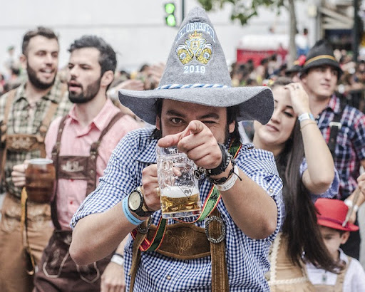 A man with a grey hat on his head and beer in his hand point at you while winking with his left eye
