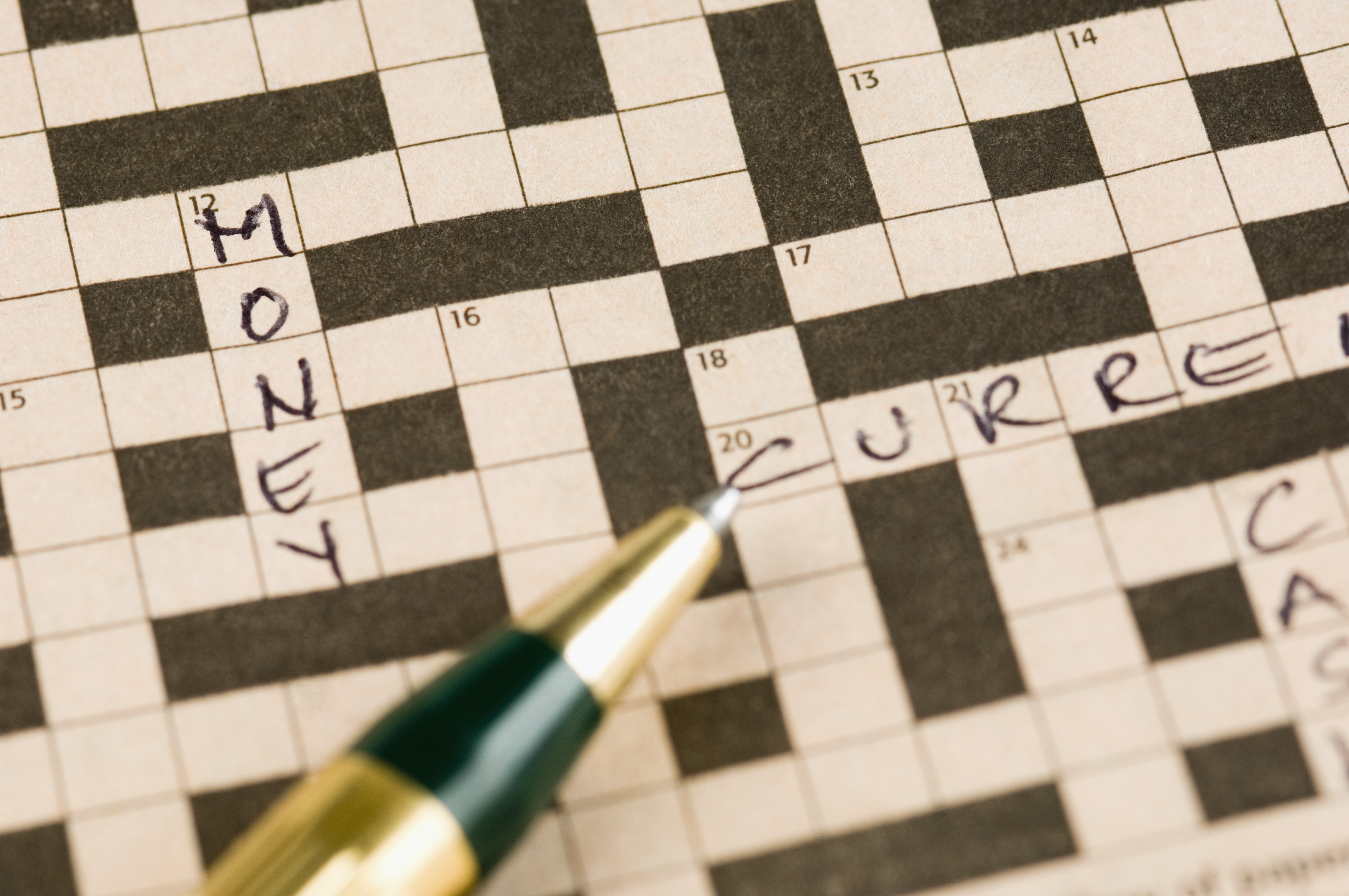 A pen poised over a crossword puzzle missing most answers aside from the word "money".