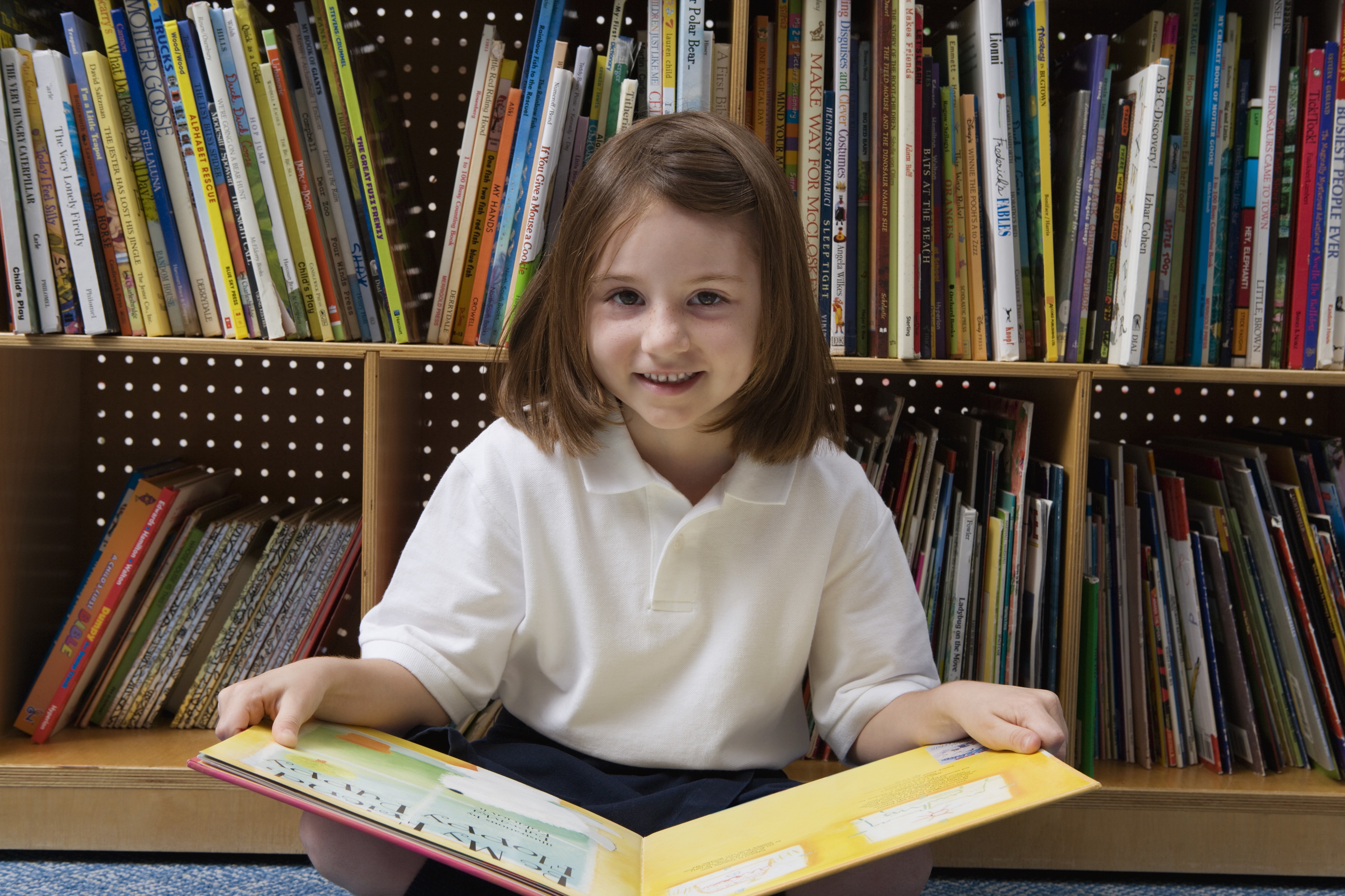Little girl sits against a bookshelf with a book in a yellow cover open on her lap